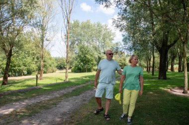Senior couple enjoying active retirement, walking together on a scenic dirt path in a lush green park, finding wellbeing and happiness outdoors during summer