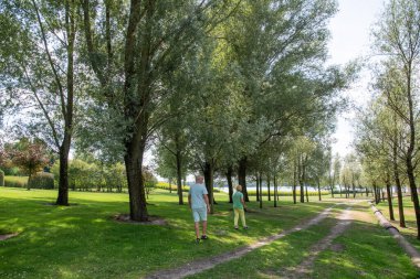 Senior couple enjoying stroll along a scenic park path, surrounded by lush green trees and views of a calm lake under the summer sun, embracing healthy active retirement