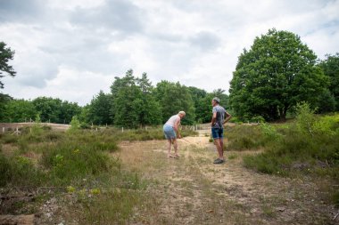 Senior couple enjoying their active retirement, walking and talking on a dirt trail through green bushes and trees under a cloudy sky, embracing healthy lifestyle and togetherness