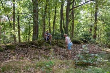 Couple taking a break during a daytime hike in a green forest, the man sitting on a moss covered bank and the woman bending over to examine the ground among the trees