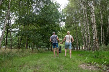 Loving couple holding hands, walking away on a green grass path in a forest of birch trees, showing togetherness, romance, and an active lifestyle outdoors with a bike