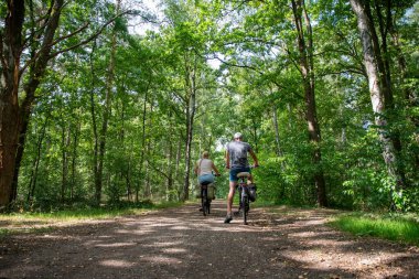 Couple cycling along a sunlit dirt path through dense green forest, enjoying summer leisure, active outdoor lifestyle and peaceful nature together on a scenic ride