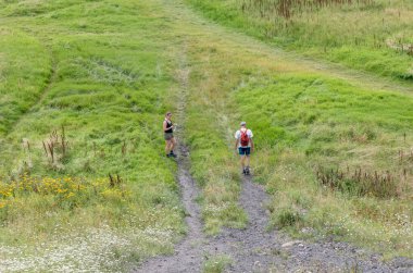 Two people walking on a winding dirt path amidst lush green grass and wildflowers, one pausing with a water bottle while the other continues the upward journey, both enjoying a warm day in nature