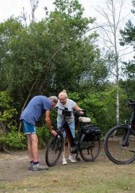 Senior man and woman standing by an electric bike, inspecting the front wheel with a caring expression, showcasing teamwork and problem solving while enjoying an active lifestyle in nature