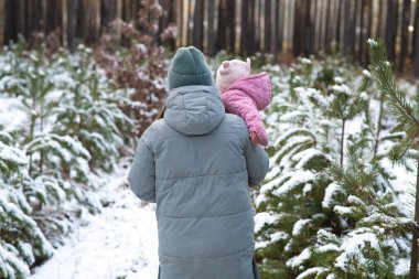 A mother in a warm winter coat carrying her child through a snowy forest path, surrounded by evergreen trees covered in snow. clipart