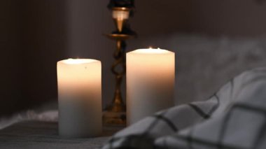 İki kalın beyaz mum, sakin ve loş bir atmosferde bir masada yavaşça yanıyor..