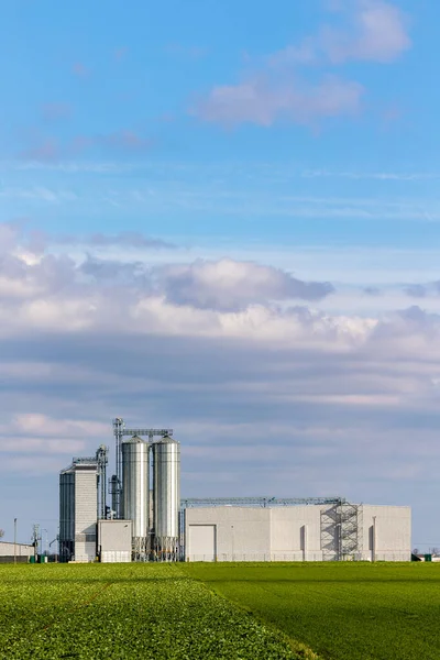 An animal feed production plant against the background of green agricultural fields. Grain silos next to the processing plant. Photo taken on a sunny day.