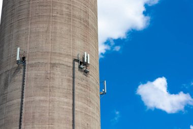 Uzun bir elektrik santrali bacasının duvarına cep telefonu antenleri monte edilmiş. Güneşli bir günde mavi gökyüzüne karşı bir nesne.