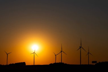 Birçok rüzgar türbini gün batımında turuncu bir gökyüzüne karşı yavaşça dönüyor. Akşamın erken saatlerinde çekilen statik çekim..      