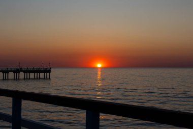 Sakin bir denizin üzerinde büyük bir turuncu güneş batıyor. Balıkçılar iskelede balık tutuyor. Bulutsuz bir akşamda çekilen statik çekim       