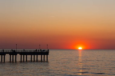 Sakin bir denizin üzerinde büyük bir turuncu güneş batıyor. Balıkçılar iskelede balık tutuyor. Bulutsuz bir akşamda çekilen statik çekim       