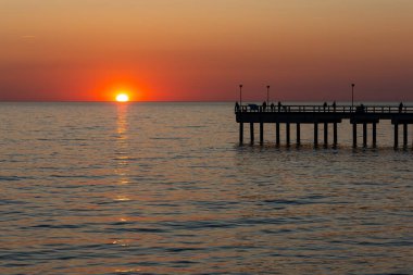 Sakin bir denizin üzerinde büyük bir turuncu güneş batıyor. Balıkçılar iskelede balık tutuyor. Bulutsuz bir akşamda çekilen statik çekim       