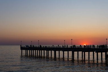 Sakin bir denizin üzerinde büyük bir turuncu güneş batıyor. Balıkçılar iskelede balık tutuyor. Bulutsuz bir akşamda çekilen statik çekim       