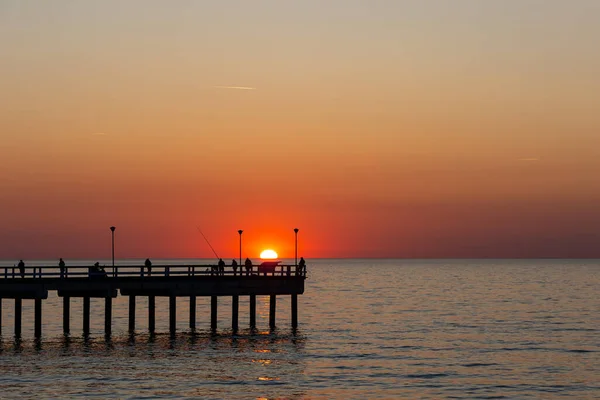 Sakin bir denizin üzerinde büyük bir turuncu güneş batıyor. Balıkçılar iskelede balık tutuyor. Bulutsuz bir akşamda çekilen statik çekim       