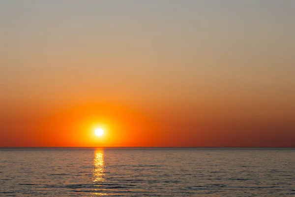 A large orange sun setting over a calm sea. Static shot taken during a cloudless evening