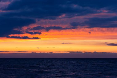 Denizin üzerinde turuncu gün batımı. Dalgalar sahile çarpıyor, dramatik bir gökyüzü. Akşamın erken saatlerinde çekilen statik bir çekim.        