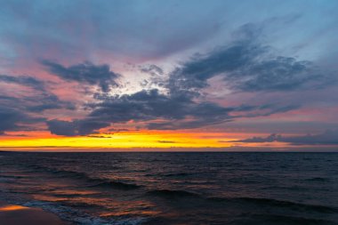 Denizin üzerinde turuncu gün batımı. Dalgalar sahile çarpıyor, dramatik bir gökyüzü. Akşamın erken saatlerinde çekilen statik bir çekim.        