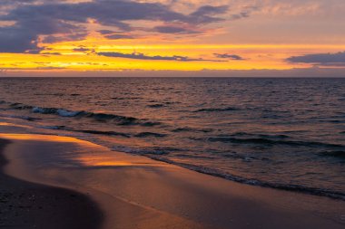 Denizin üzerinde turuncu gün batımı. Dalgalar sahile çarpıyor, dramatik bir gökyüzü. Akşamın erken saatlerinde çekilen statik bir çekim.        