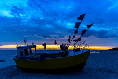 Sarı turuncu bir balıkçı teknesi gün batımında sahile yanaştı. Bulutlu dramatik günbatımı gökyüzü. Fotoğraf akşamları çok az ışıklandırmayla çekiliyor.
