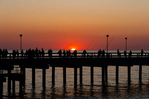 Dark outlines of people walking on the sea pier during the setting sun. The sky lit orange with the last rays of the sun.