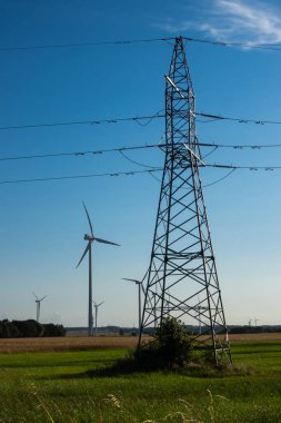 Rüzgar türbinleri ve mavi gökyüzüne karşı yüksek voltajlı elektrik pilonu. Türbinlerin arkasında güneş parlıyor.