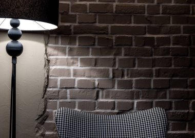 Gri, rahat, houndstooth desenli bir koltuk gri tuğla bir duvarın karşısında duruyor. Sahne, bir lambanın atmosferik ışığıyla aydınlatılır..
