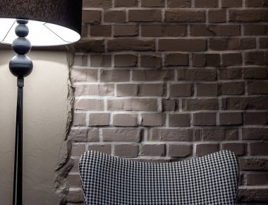 Gri, rahat, houndstooth desenli bir koltuk gri tuğla bir duvarın karşısında duruyor. Sahne, bir lambanın atmosferik ışığıyla aydınlatılır..