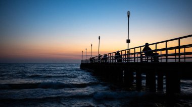 Silhouetts of a people sitting on the benches at the sea pier at sunset. The natural glow of the orange and navy blue sky at the evening