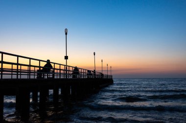Silhouetts of a people sitting on the benches at the sea pier at sunset. The natural glow of the orange and navy blue sky at the evening