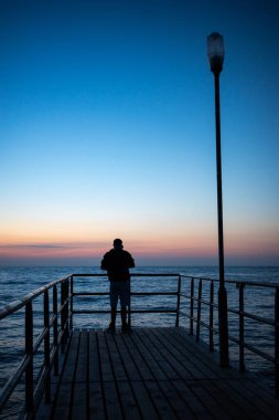 Bir adamın silueti gün batımında bir rıhtımın ucunda duruyor. Akşamları turuncu ve lacivert gökyüzünün parlaklığı
