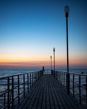 Bir adamın silueti gün batımında bir rıhtımın ucunda duruyor. Akşamları turuncu ve lacivert gökyüzünün parlaklığı