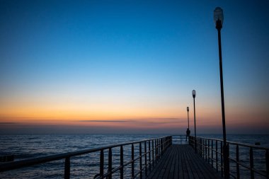Bir adamın silueti gün batımında bir rıhtımın ucunda duruyor. Akşamları turuncu ve lacivert gökyüzünün parlaklığı