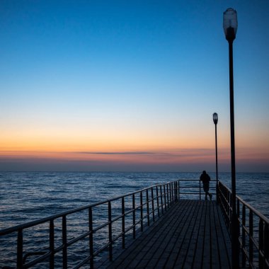 Bir adamın silueti gün batımında bir rıhtımın ucunda duruyor. Akşamları turuncu ve lacivert gökyüzünün parlaklığı