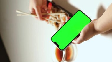Evde suşi yerken akıllı telefon kullanan bir kadın. Yeşil ekranlı akıllı telefon. Restoranlar, yemek dağıtımı, yemek siteleri ve uygulamalar için kopyalama alanı