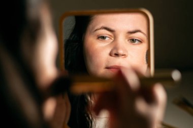 Evde makyaj yapan bir kadın aynaya bakıyor. Fırçayla makyaj yapıyorum. Yüzüne cilt pudrası sürüyorum. Kırmızı noktaları kaplıyor. Aynadaki yansıma.