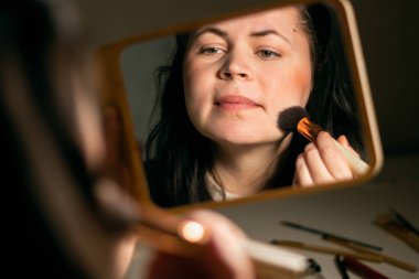 Evde makyaj yapan bir kadın aynaya bakıyor. Fırçayla makyaj yapıyorum. Yüzüne cilt pudrası sürüyorum. Kırmızı noktaları kaplıyor. Aynadaki yansıma.