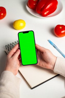 Defterde sağlıklı beslenme planı hazırlayan bir kadın. Beslenme planı. Telefondaki yeşil ekran, online kilo verme programı için hazırlanıyor. Defter, sebze ve telefon düzlüğü.