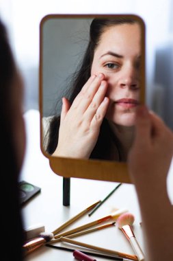 Millennial Kafkas kadınının aynaya bakarken makyaj yaparken yüz kremi sürüşünün dikey görüntüsü. Aynadaki yansıma. Hazırlanıyor. Güzellik konsepti.