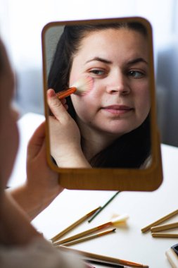 Millennial Kafkas kadınının aynaya bakarken yanaklarına makyaj fırçasıyla allık sürerken görüntüsü. Aynadaki yansıma. Hazırlanıyor. Güzellik konsepti.