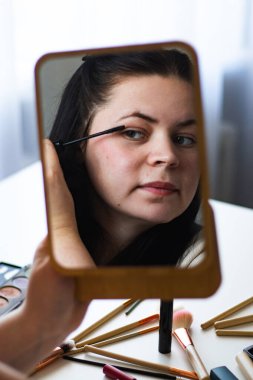 Millennial Kafkas kadınının dikey görüntüsü aynaya bakarken maskara sürerek makyaj yapıyor. Aynadaki yansıma. Hazırlanıyor. Güzellik konsepti.