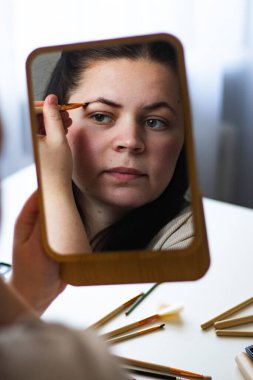 Millennial Kafkas kadınının dikey görüntüsü aynaya bakarak kaşlarını boyuyor. Aynadaki yansıma. Hazırlanıyor. Güzellik konsepti.