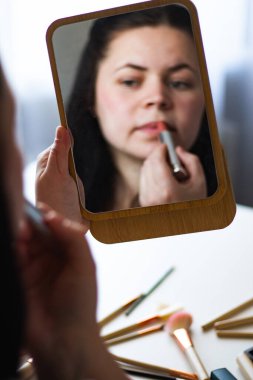 Bin yıllık beyaz bir kadının aynaya bakarken dudaklarına ruj sürerek makyaj yaptığı dikey bir görüntü. Aynadaki yansıma. Hazırlanıyor. Güzellik konsepti.