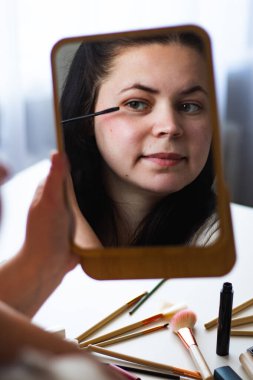 Millennial Kafkas kadınının dikey görüntüsü aynaya bakarken maskara sürerek makyaj yapıyor. Aynadaki yansıma. Hazırlanıyor. Güzellik konsepti.