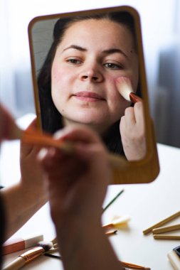 Millennial Kafkas kadınının aynaya bakarken yanaklarına makyaj fırçasıyla allık sürerken görüntüsü. Aynadaki yansıma. Hazırlanıyor. Güzellik konsepti.