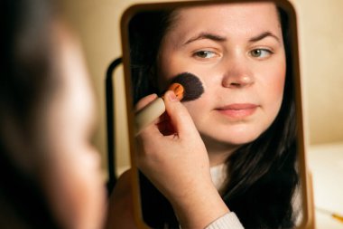 Evde makyaj yapan bir kadın aynaya bakıyor. Fırçayla makyaj yapıyorum. Yüze allık sürüyorum. Aynadaki yansıma.