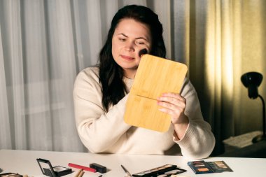 Evde makyaj yapan bir kadın aynaya bakıyor. Fırçayla makyaj yapıyorum. Yüze allık sürüyorum. Aynadaki yansıma.