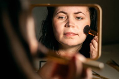 Evde makyaj yapan bir kadın aynaya bakıyor. Fırçayla makyaj yapıyorum. Yüzüne cilt pudrası sürüyorum. Kırmızı noktaları kaplıyor. Aynadaki yansıma.