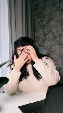 Evde dizüstü bilgisayarla çalışan yorgun bir kadın elinde gözlüklerle gözlerini ovuşturuyor ve yorgun hissediyor. Stres ve hüsran konsepti. Yorgun gözler.