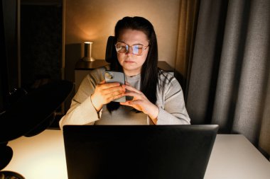 Dizüstü bilgisayarla çalışırken akıllı telefon kullanan gözlüklü ciddi bir serbest kadın.