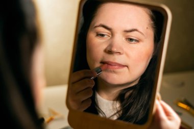 Aynaya bakan ve dudaklarına ruj süren bir kadın. Makyaj. Etkinlik için hazırlanılıyor. Güzellik ve kişisel bakım konsepti. Aynadaki yansıma